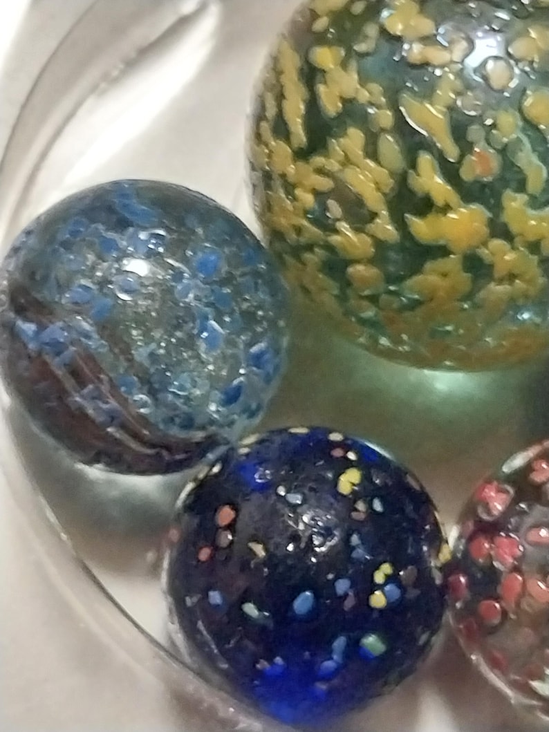 Op de afbeelding: Een verzameling van vier glazen knikkers met verschillende patronen. E&eacute;n knikker is blauw met witte stippen, een andere is groen met gele stippen, een andere is blauw met veelkleurige stippen en de laatste is helder met rode en witte stippen.