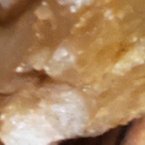 May include: Close-up of a rough, textured, light brown surface with white and tan highlights. The surface appears to be a natural material, possibly a rock or mineral, with a slightly uneven texture.