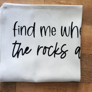 May include: A folded white canvas cloth with the black text "find me where the rocks are". The cloth has a stitched edge and is resting on a wooden surface.