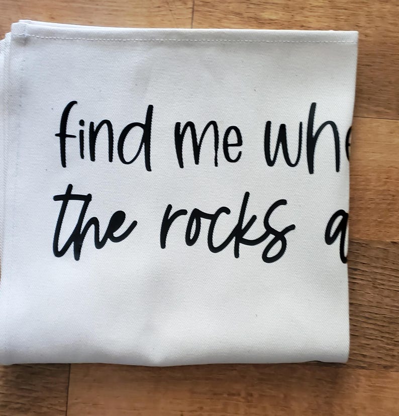 May include: A folded white canvas cloth with the black text "find me where the rocks are". The cloth has a stitched edge and is resting on a wooden surface.