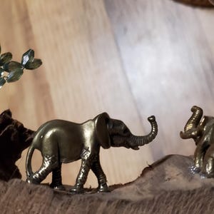May include: Two small, gold-colored elephant figurines on a wooden base. One elephant is walking, the other is standing. A small, green, crystal-like tree is also present.