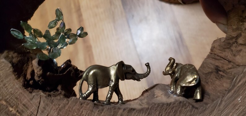 May include: Two small, gold-colored elephant figurines on a wooden base. One elephant is walking, the other is standing. A small, green, crystal-like tree is also present.