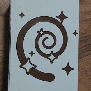May include: A rectangular, off-white box featuring a brown spiral design with star accents. The spiral is centered, with four stars surrounding it and two additional stars above. The box is set against a wooden surface.
