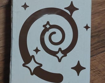 May include: A rectangular, off-white box featuring a brown spiral design with star accents. The spiral is centered, with four stars surrounding it and two additional stars above. The box is set against a wooden surface.
