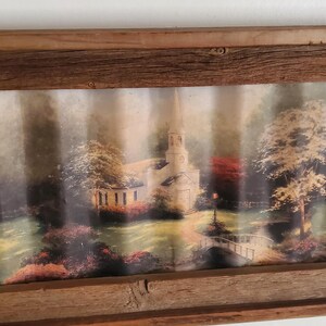 May include: A framed landscape print featuring a church and trees in a rustic wooden frame. The artwork depicts a serene scene with a bridge, lush greenery, and a soft, diffused light, creating a tranquil atmosphere.