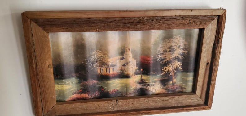 May include: A framed landscape print featuring a church and trees in a rustic wooden frame. The artwork depicts a serene scene with a bridge, lush greenery, and a soft, diffused light, creating a tranquil atmosphere.