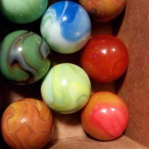 May include: A collection of colorful marbles in a cardboard box. The marbles are various colors, including green, blue, orange, and red, with swirled patterns. The marbles are round and smooth, and the box is brown.