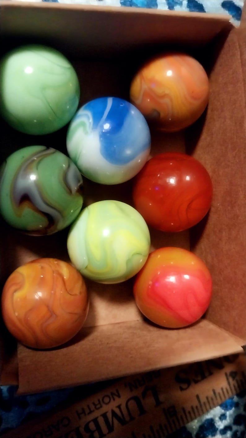 May include: A collection of colorful marbles in a cardboard box. The marbles are various colors, including green, blue, orange, and red, with swirled patterns. The marbles are round and smooth, and the box is brown.
