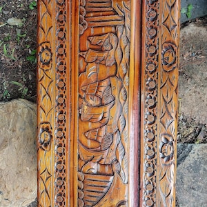 May include: A rectangular wooden panel with intricate carvings. The central panel features a detailed relief of sailing ships and a mountain scene. The border is adorned with floral and leaf patterns. The wood has a warm, reddish-brown hue.