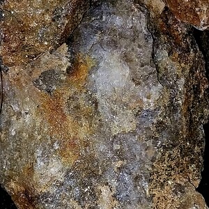 May include: Close-up of a rough, textured rock formation. The stone displays a mix of colors, including shades of brown, gold, gray, and white. The surface has a crystalline appearance with areas of sparkle and shine, suggesting mineral deposits.