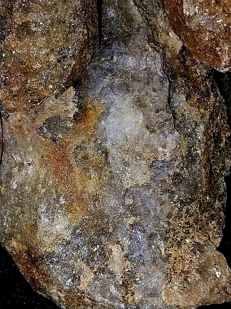 May include: Close-up of a rough, textured rock formation. The stone displays a mix of colors, including shades of brown, gold, gray, and white. The surface has a crystalline appearance with areas of sparkle and shine, suggesting mineral deposits.