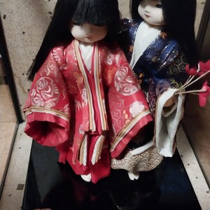 May include: Two Japanese dolls with black hair and traditional clothing. One doll wears a red kimono with gold accents, the other a blue kimono. They stand on a black base, showcasing detailed craftsmanship.