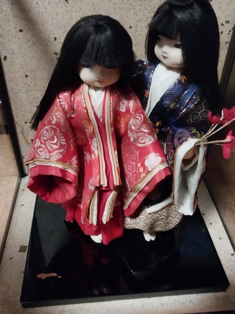 May include: Two Japanese dolls with black hair and traditional clothing. One doll wears a red kimono with gold accents, the other a blue kimono. They stand on a black base, showcasing detailed craftsmanship.