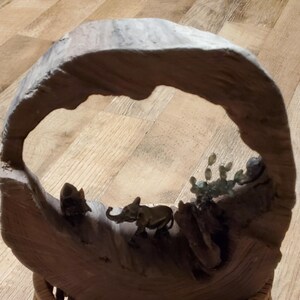 May include: A wooden sculpture featuring an elephant and other animal figures within a natural arch. The sculpture is made of light brown wood and has a woven base. The elephant is small and detailed.
