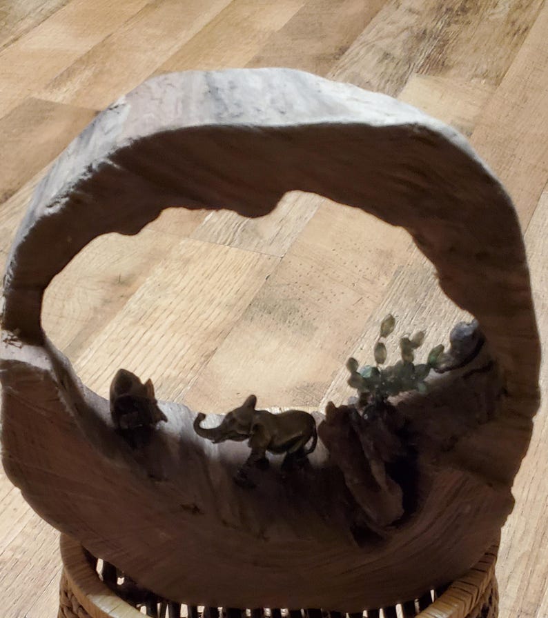 May include: A wooden sculpture featuring an elephant and other animal figures within a natural arch. The sculpture is made of light brown wood and has a woven base. The elephant is small and detailed.