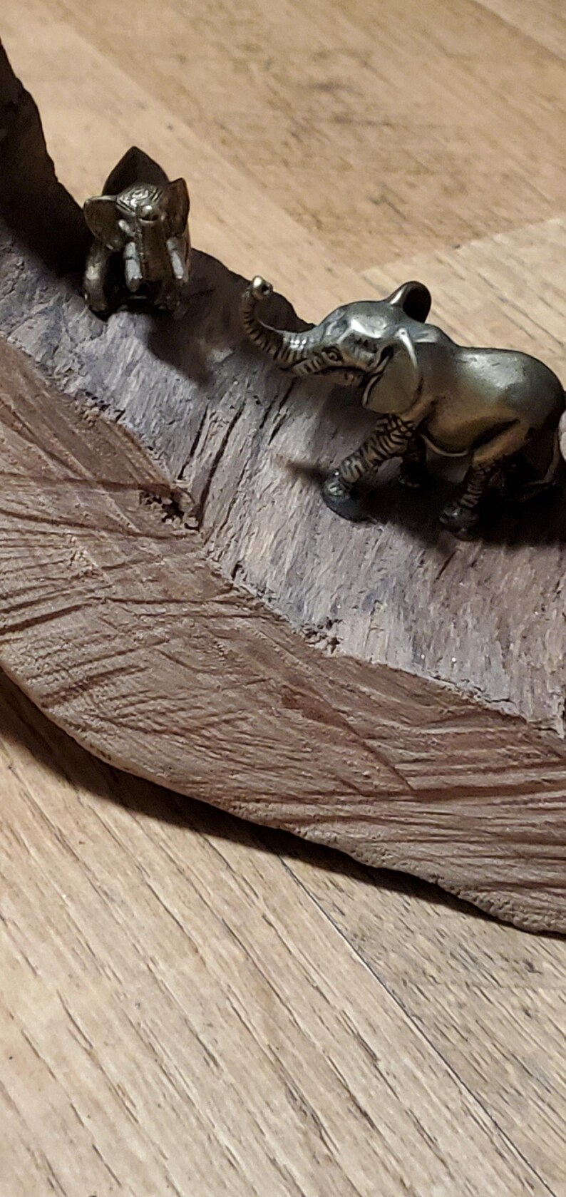 May include: Two small, bronze-colored elephant figurines on a piece of wood. One elephant is standing upright, while the other is in a walking pose. The wood has a textured, brown surface, and the background is a light-colored wooden floor.