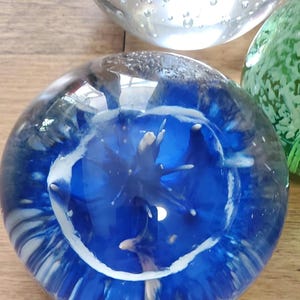 May include: A clear glass paperweight with a striking blue interior design and white accents. The design has an abstract shape. Other glass spheres are visible in the background. The paperweight is a decorative item.