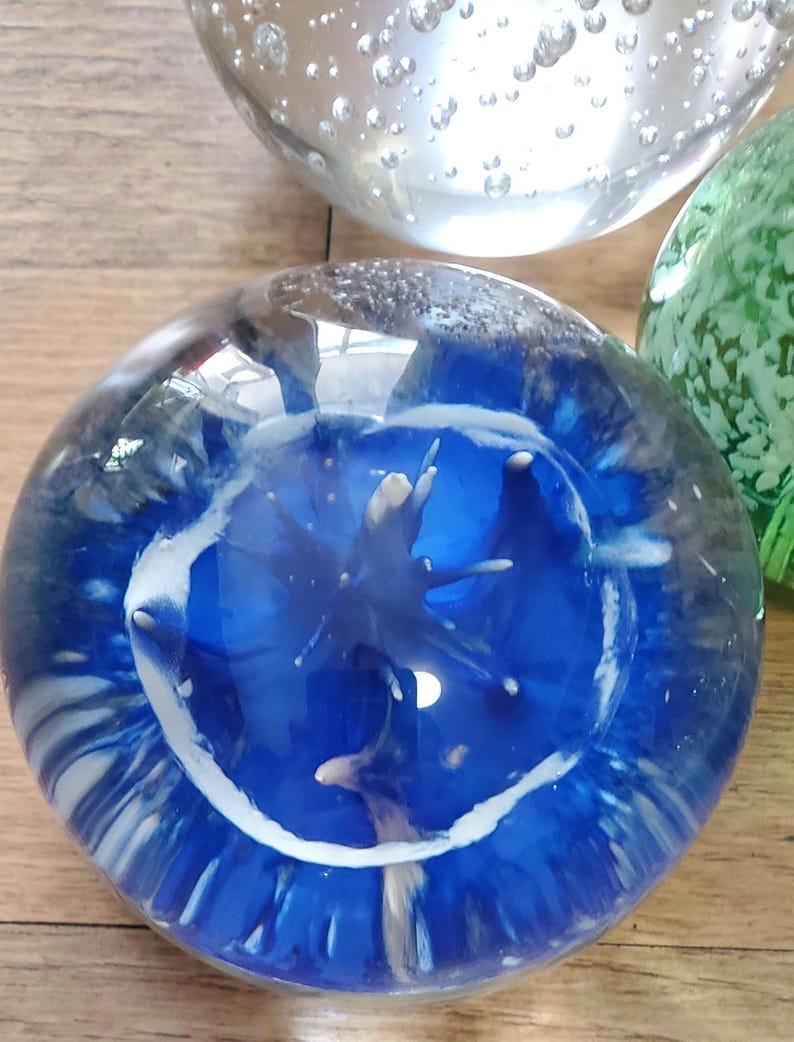May include: A clear glass paperweight with a striking blue interior design and white accents. The design has an abstract shape. Other glass spheres are visible in the background. The paperweight is a decorative item.