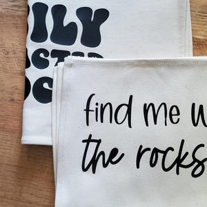 May include: Two off-white canvas tea towels with black text. One towel reads "ILY" and "STAY DOPE". The other towel says "find me with the rocks". The towels are folded and stacked on a wooden surface.