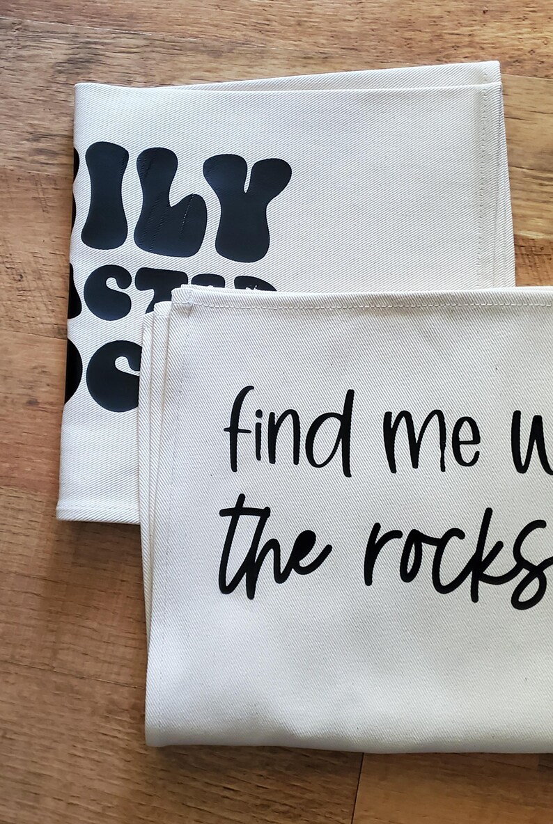 May include: Two off-white canvas tea towels with black text. One towel reads "ILY" and "STAY DOPE". The other towel says "find me with the rocks". The towels are folded and stacked on a wooden surface.