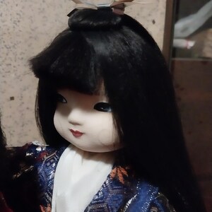 May include: A Japanese doll with long black hair and a serene expression. The doll wears a blue and gold patterned kimono with a white under-layer. A decorative hairpiece adorns the top of the head. The doll's face is painted with delicate features.