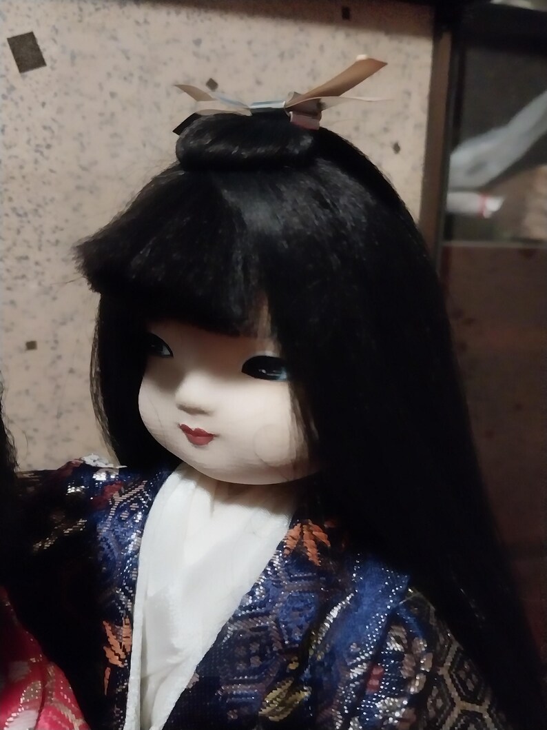 May include: A Japanese doll with long black hair and a serene expression. The doll wears a blue and gold patterned kimono with a white under-layer. A decorative hairpiece adorns the top of the head. The doll's face is painted with delicate features.