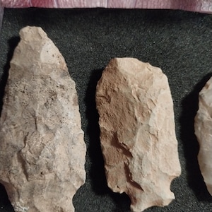 May include: Three stone arrowheads, likely used by Native Americans, are displayed on a black background. The arrowheads are made of a light brown stone with a rough texture.