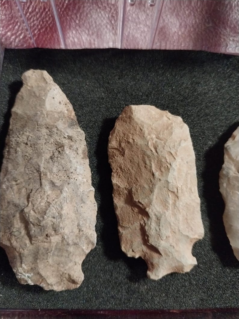 May include: Three stone arrowheads, likely used by Native Americans, are displayed on a black background. The arrowheads are made of a light brown stone with a rough texture.