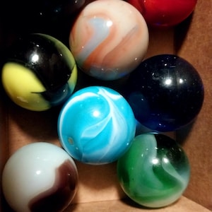 May include: A collection of colorful marbles in a cardboard box. The marbles are various colors, including black, red, blue, green, and white, with swirling patterns. The marbles are a classic toy.
