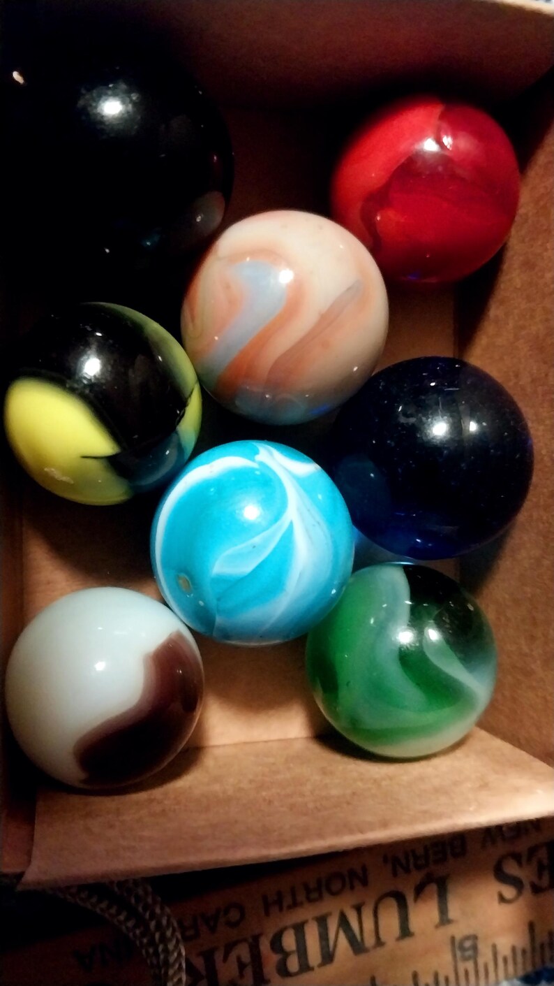 May include: A collection of colorful marbles in a cardboard box. The marbles are various colors, including black, red, blue, green, and white, with swirling patterns. The marbles are a classic toy.