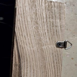 May include: A light brown wooden plaque with a natural wood grain pattern. The plaque has a metal hook attached to the top right corner.