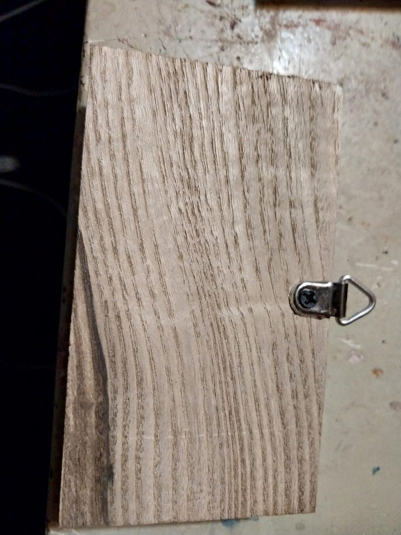 May include: A light brown wooden plaque with a natural wood grain pattern. The plaque has a metal hook attached to the top right corner.