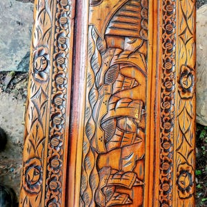 May include: A detailed close-up of a wooden carved panel. The panel features intricate relief carvings of floral patterns and a central scene with architectural elements. The wood has a warm, reddish-brown hue, with visible grain and texture.