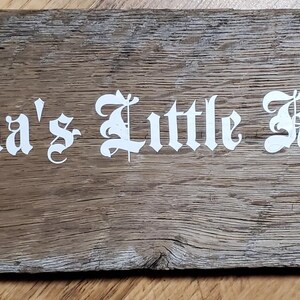May include: A rectangular wooden sign with the text "Karma's Little Helper" in white gothic lettering. The wood has a natural, weathered appearance with visible grain and knots. The sign has a rustic, handmade aesthetic.