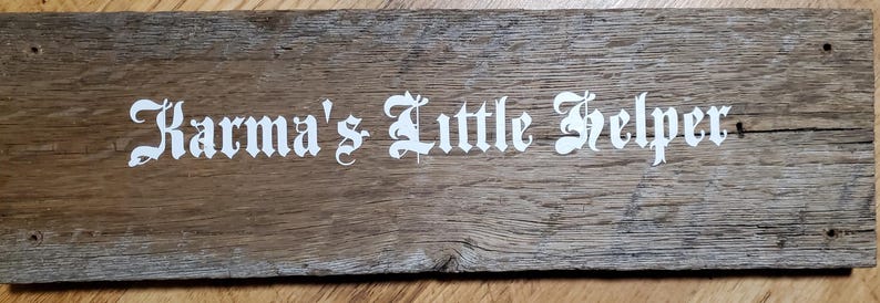 May include: A rectangular wooden sign with the text "Karma's Little Helper" in white gothic lettering. The wood has a natural, weathered appearance with visible grain and knots. The sign has a rustic, handmade aesthetic.