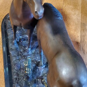 May include: Bronze sculpture of two horses. One horse stands upright, while the other leans in, touching the first horse. The sculpture is set on a black rectangular base, with detailed textures and colors.