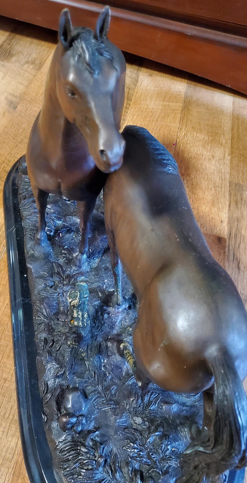 May include: Bronze sculpture of two horses. One horse stands upright, while the other leans in, touching the first horse. The sculpture is set on a black rectangular base, with detailed textures and colors.