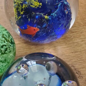 May include: Glass paperweights with underwater designs. One has a blue and yellow coral reef with orange fish. Another features blue and white swirls with bubbles. Decorative glass art pieces.