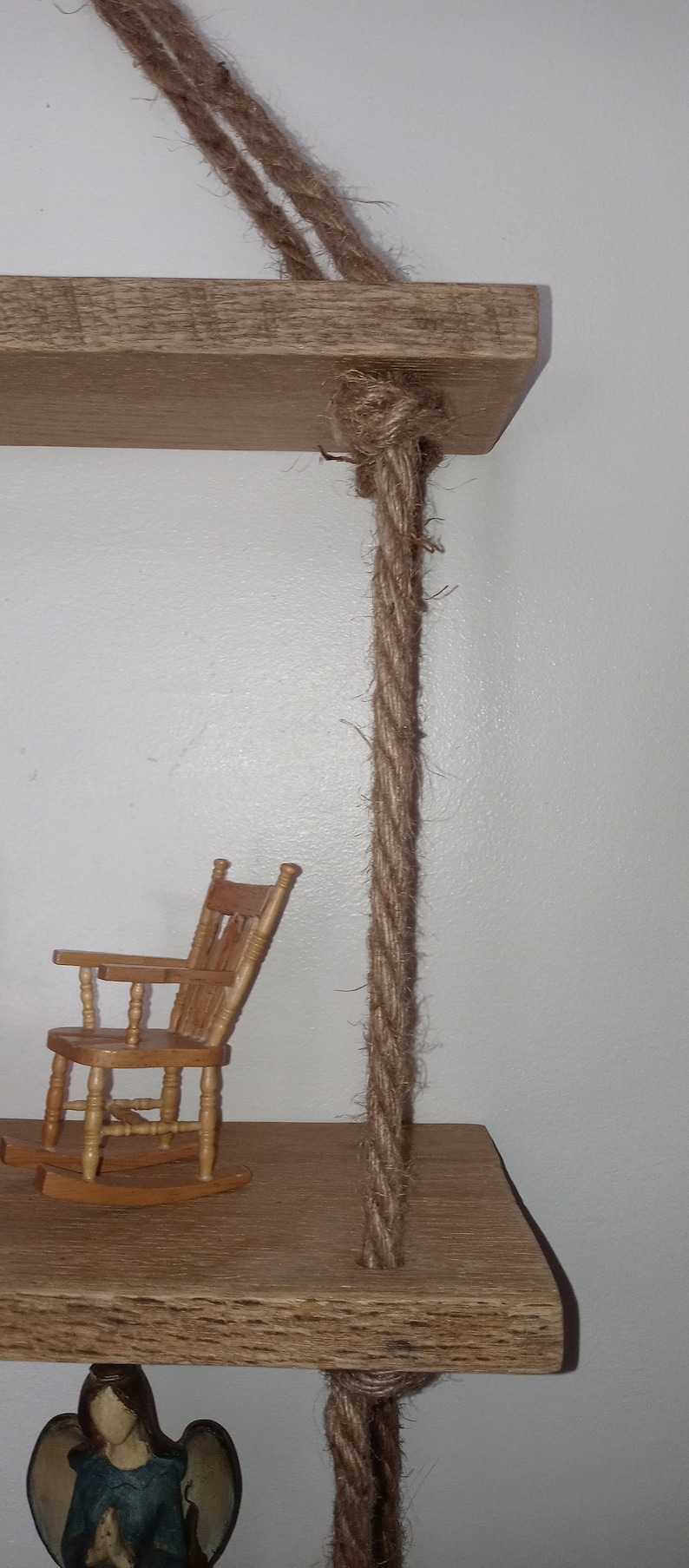 May include: Two wooden shelves hanging from the ceiling with thick brown rope. The top shelf has a small wooden rocking chair on it. The bottom shelf has a small angel figurine on it.