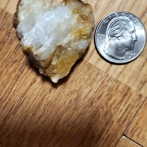 May include: A heart-shaped, rough-textured stone with white and yellow hues, next to a U.S. quarter coin dated 2024. The stone is on a wooden surface.