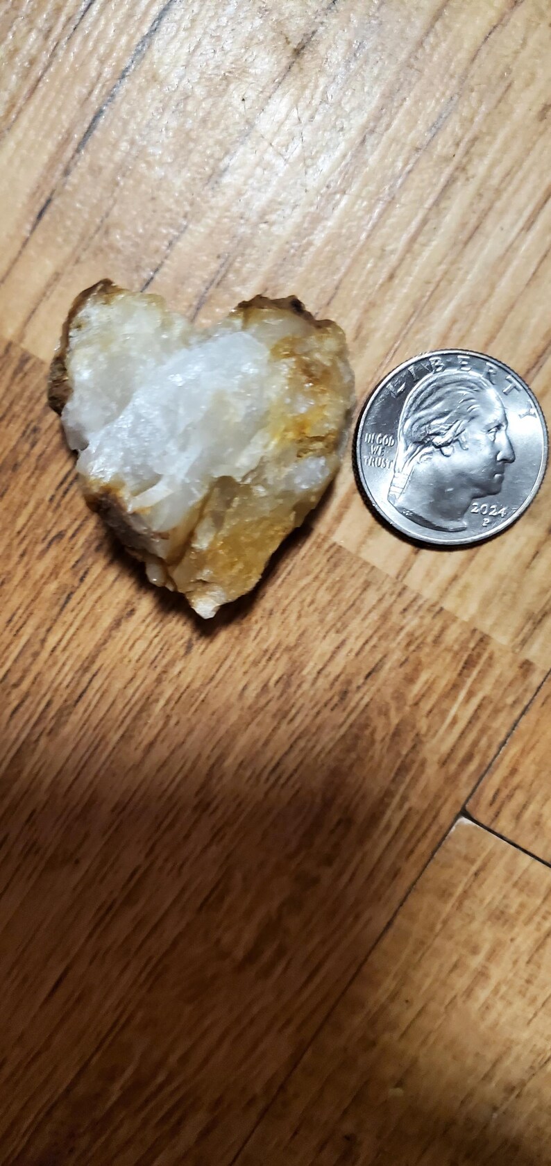 May include: A heart-shaped, rough-textured stone with white and yellow hues, next to a U.S. quarter coin dated 2024. The stone is on a wooden surface.