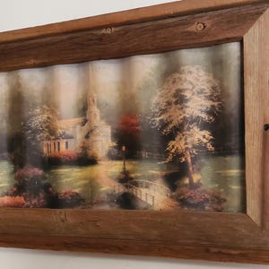 May include: A framed landscape print featuring a church, trees, and a bridge. The artwork is set within a rustic, rectangular wooden frame. The image has a soft, muted color palette with shades of green, brown, and beige.