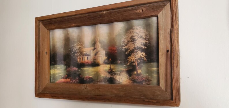 May include: A framed landscape print featuring a church, trees, and a bridge. The artwork is set within a rustic, rectangular wooden frame. The image has a soft, muted color palette with shades of green, brown, and beige.