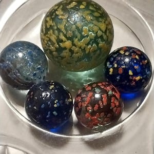 Op de afbeelding: Vijf glazen knikkers met verschillende kleurpatronen. De knikkers zijn blauw, groen, rood en zwart met witte stippen. De knikkers bevinden zich in een glazen kom.
