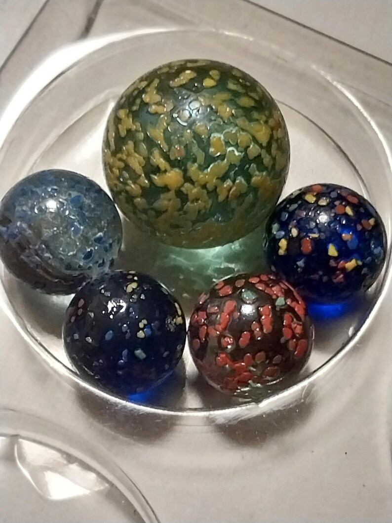 Op de afbeelding: Vijf glazen knikkers met verschillende kleurpatronen. De knikkers zijn blauw, groen, rood en zwart met witte stippen. De knikkers bevinden zich in een glazen kom.