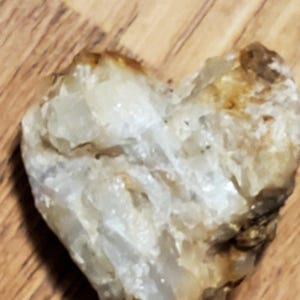 May include: A heart-shaped, natural stone with a rough, textured surface. The stone is primarily off-white with areas of tan and brown, resting on a wooden surface.