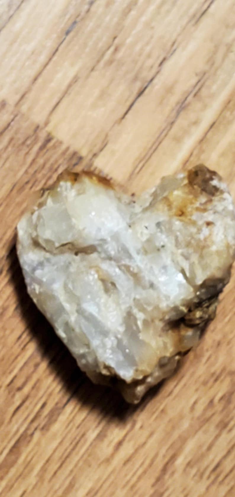 May include: A heart-shaped, natural stone with a rough, textured surface. The stone is primarily off-white with areas of tan and brown, resting on a wooden surface.