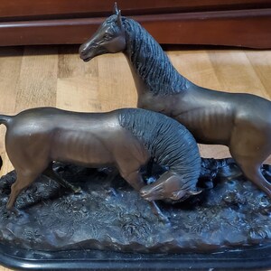 May include: Bronze sculpture of two horses. One horse stands tall, while the other lowers its head to graze. The sculpture is mounted on a black, oval base, and the horses are depicted with detailed musculature.