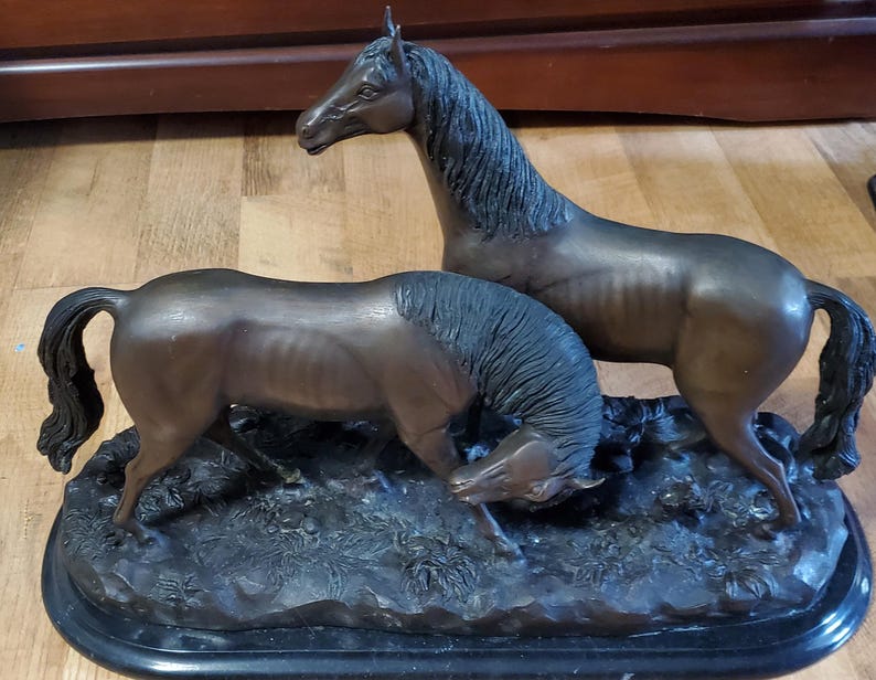 May include: Bronze sculpture of two horses. One horse stands tall, while the other lowers its head to graze. The sculpture is mounted on a black, oval base, and the horses are depicted with detailed musculature.