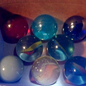 May include: A collection of colorful glass marbles in various shades of blue, red, and clear. Some marbles have internal swirls of color. The marbles are in a cardboard box with a patterned edge.
